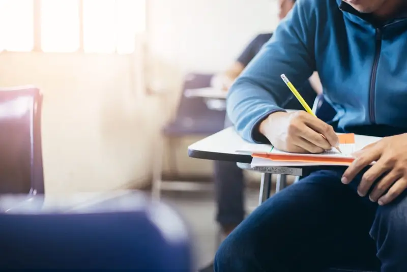 Someone sits at a single seat desk completing an exam with a pencil.