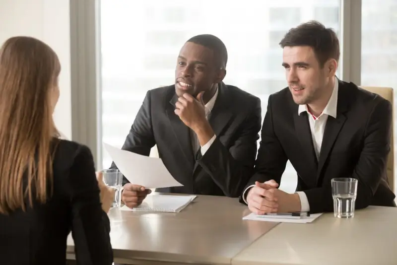 Two people in suits sit across a table from a third person who they are interviewing.