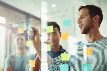 A team of three stands on the other side of a transparent board covered in post-it notes.