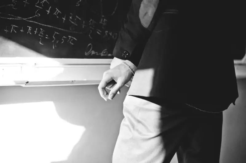 A man stands thoughtfully in front of a chalkboard covered in mathematical symbols he is working on.